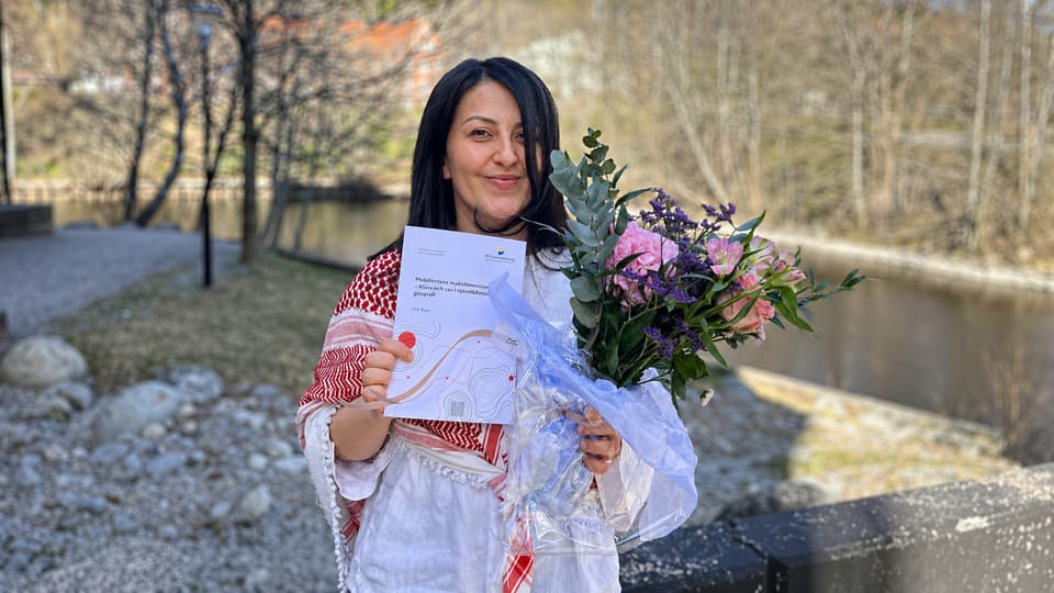 Kvinna står utomhus hållandes blommor och ett diplom.
