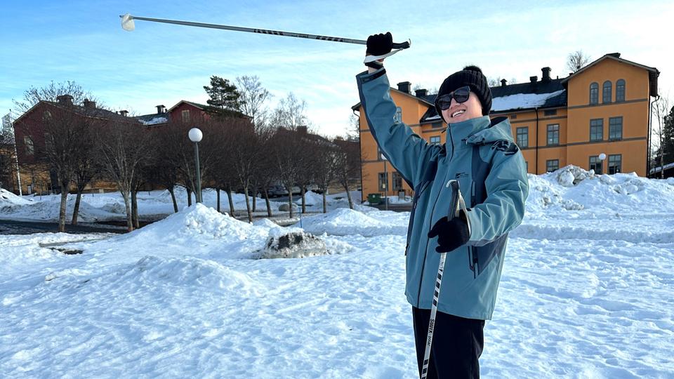 Ung man med stavar i händerna står på en snötäckt plan och sträcker upp ena handen i luften och ler