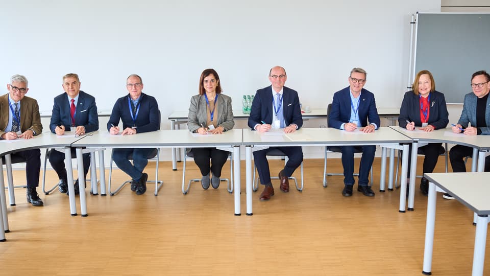 8 personer i kostym sitter vid bord i en halvcirkel