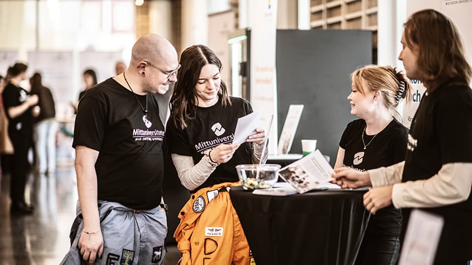 Two male and two female students in black Mid Sweden University t-shirts stand around a table reading on a postcard.