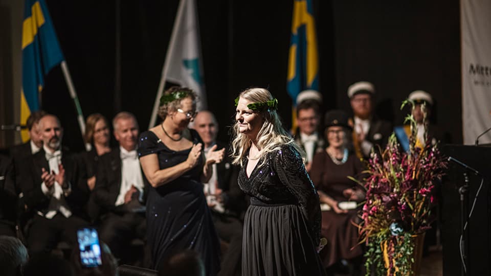 A well-dressed woman with a laurel wreath stands on a stage. 
