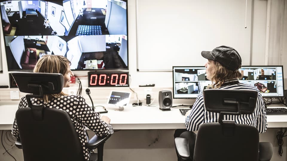Two people are sitting at a long work table. In front of them are several computer screens, a microphone and other technical equipment.