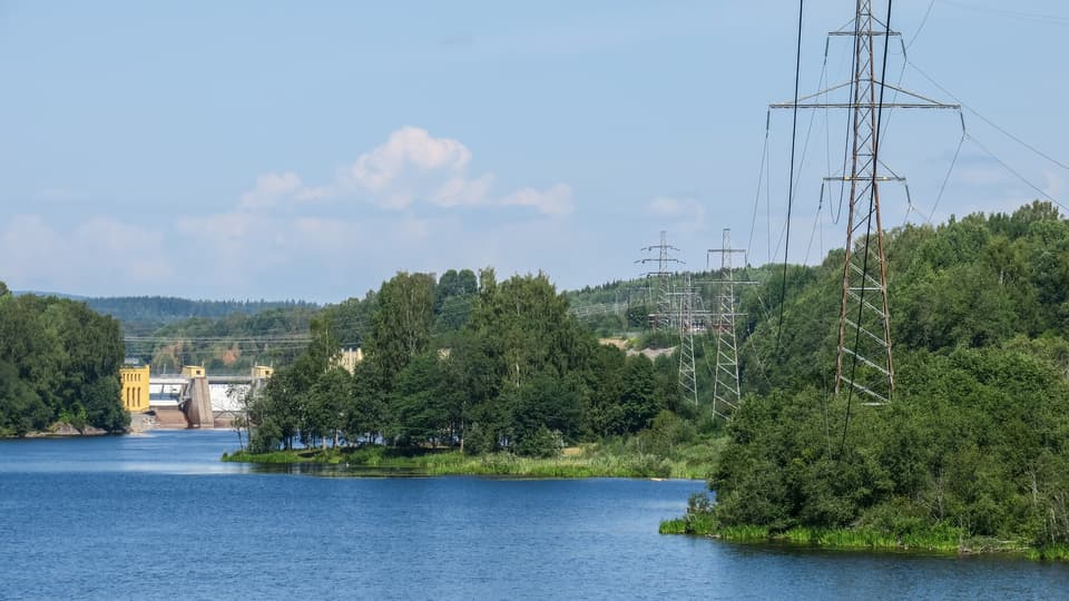  Electricity poles stand in a landscape with forest and a watercourse with a hydroelectric plant visible in the background.