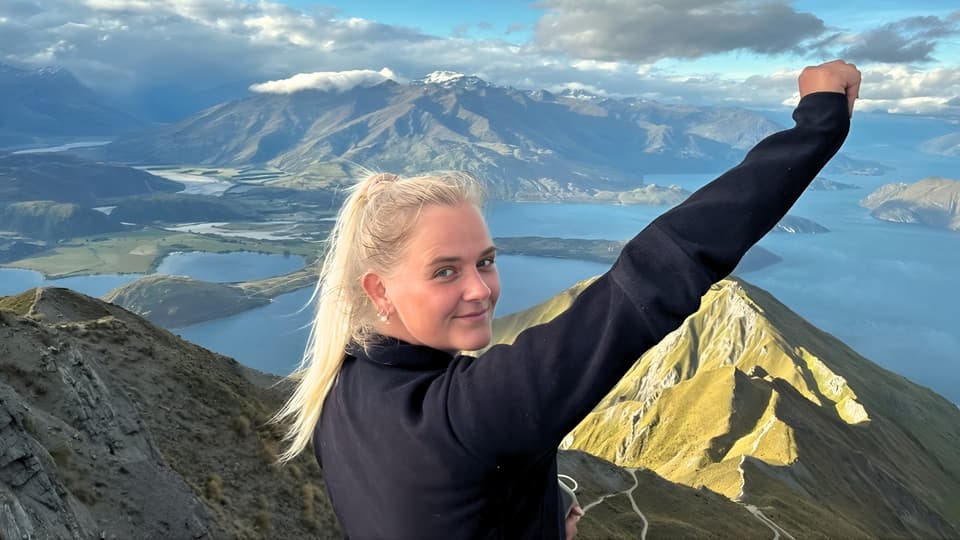 Ung kvinna står på ett berg med utsikt över havet och ett vulkanlandskap. Hon är glad och sträcker upp sin arm. 