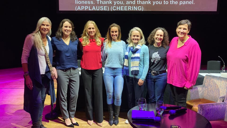 Seven women on a conference stage in front of a screen.