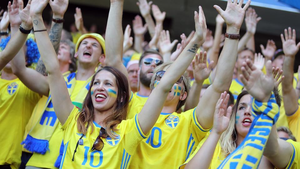 Glada människor klädda i fotbollströjor och halsdukar hejandes på Sverige på en arena. Vissa har händerna uppsträckta och vissa klappar händerna. 