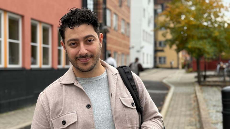 En man tittar in i kameran och ler. I bakgrunden syns några husfasader och träd.