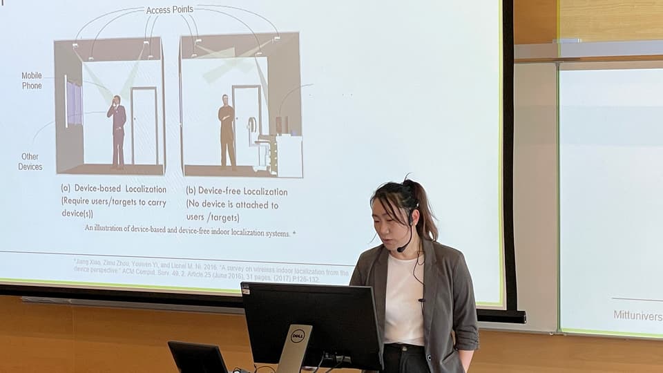 A woman with dark hair in a ponytail is standing at a computer giving a presentation.