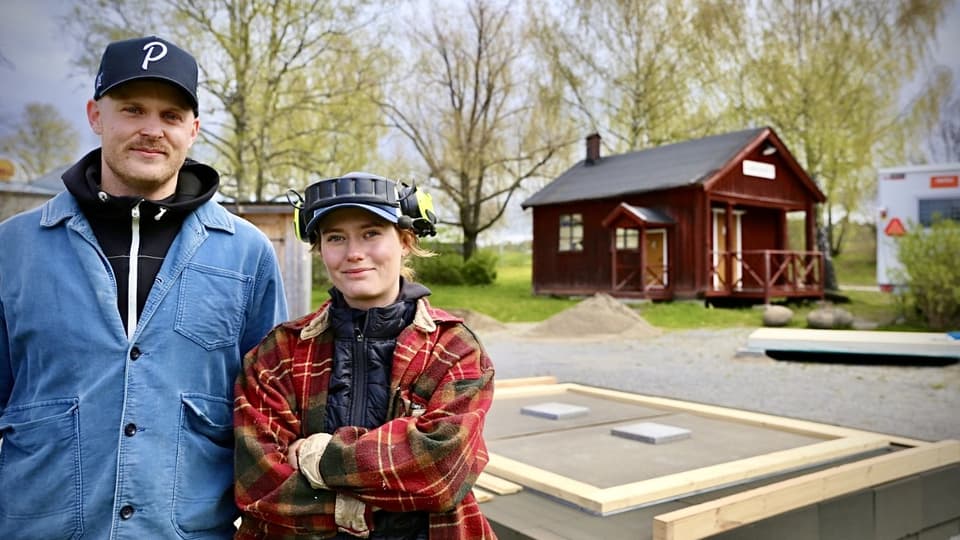 Two persons, a man and a woman, is standing togehter outside. Behind them ypu can see two small house grounds.