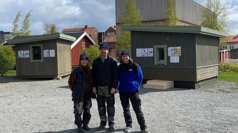 Tre personer står utomhus framför två små byggnader.