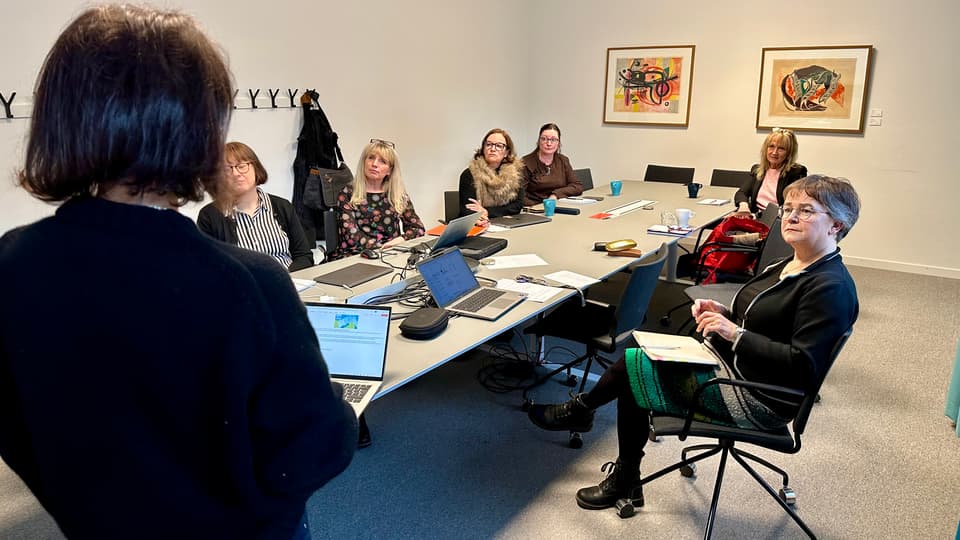 Six women are sitting around a conference table, looking at another woman who is standing with her back to the camera.