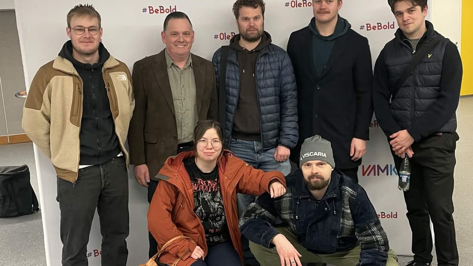 Sju personer poserar för en gruppbild framför en vit bakgrund med VAMK‑logotyp. Fem står upp och två sitter på huk framför dem. Alla bär vardagliga ytterkläder i en inomhusmiljö.