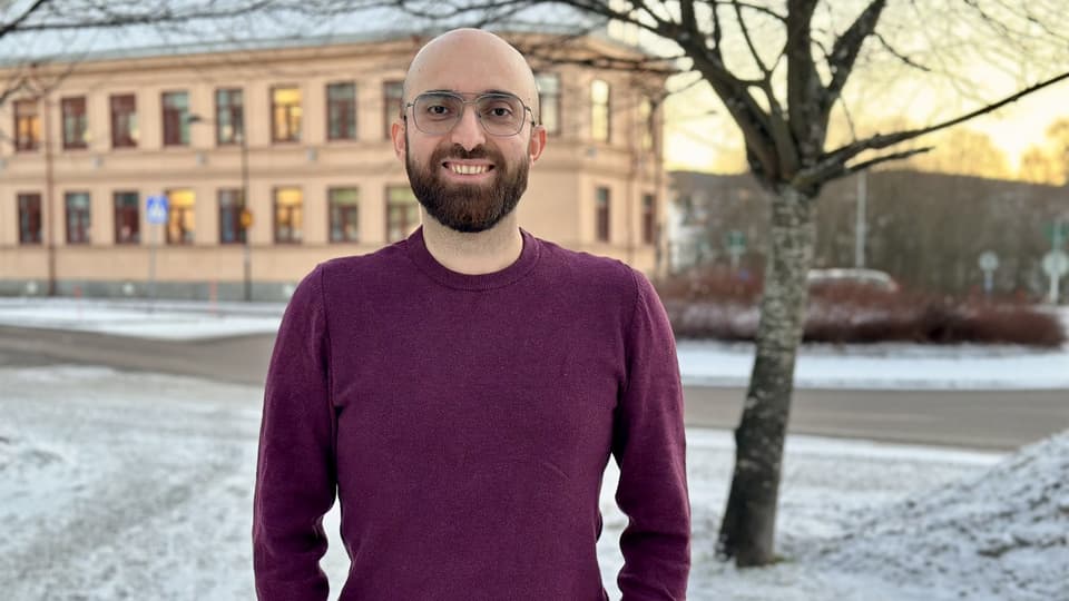 Smiling man with a shaved head, brown beard, and glasses. He is wearing a burgundy sweater and standing outdoors. The season is winter.