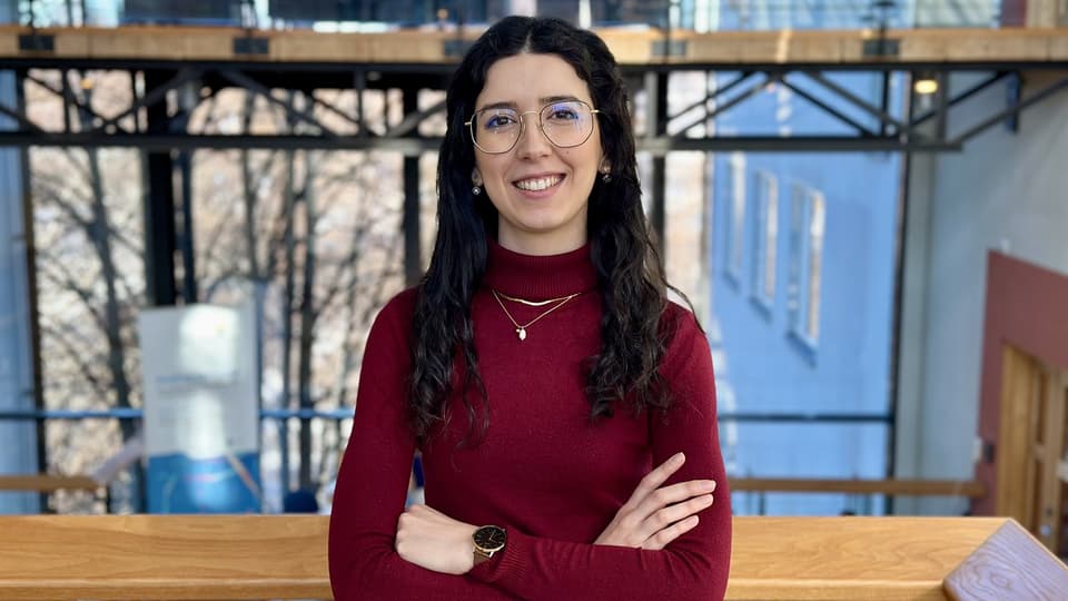 Smiling woman with dark curly hair, glasses, and a burgundy sweater standing with her arms crossed.