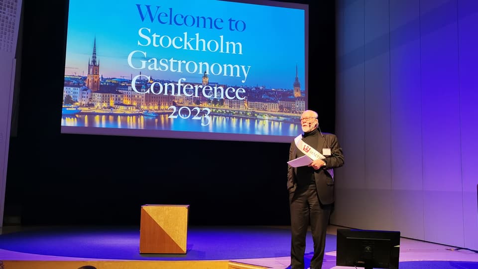 Man in a suit stands on a stage with a paper in his hand and a screen behind him with the text Stockholm Gastronomy Conference 2023