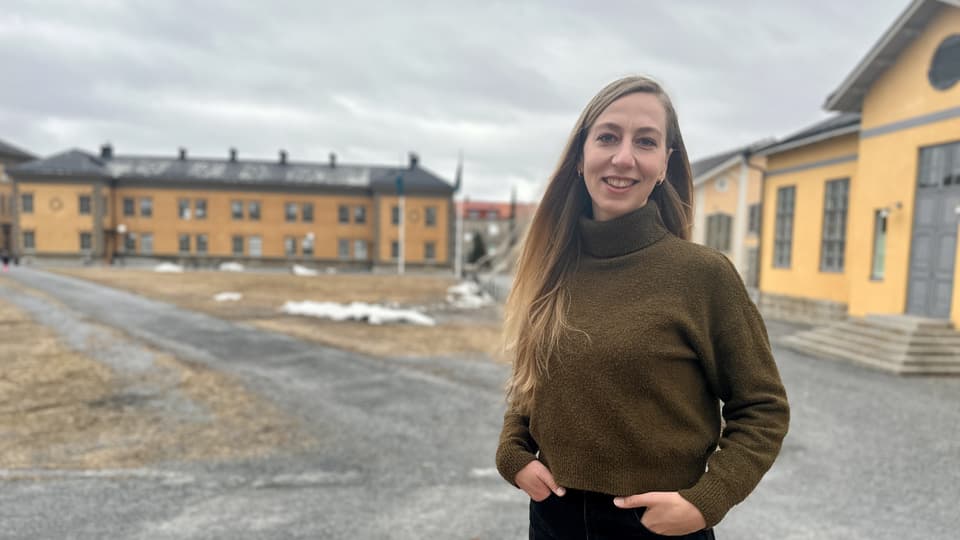 Woman in brown polo shirt and black trousers standing outdoors in front of large yellow buildings