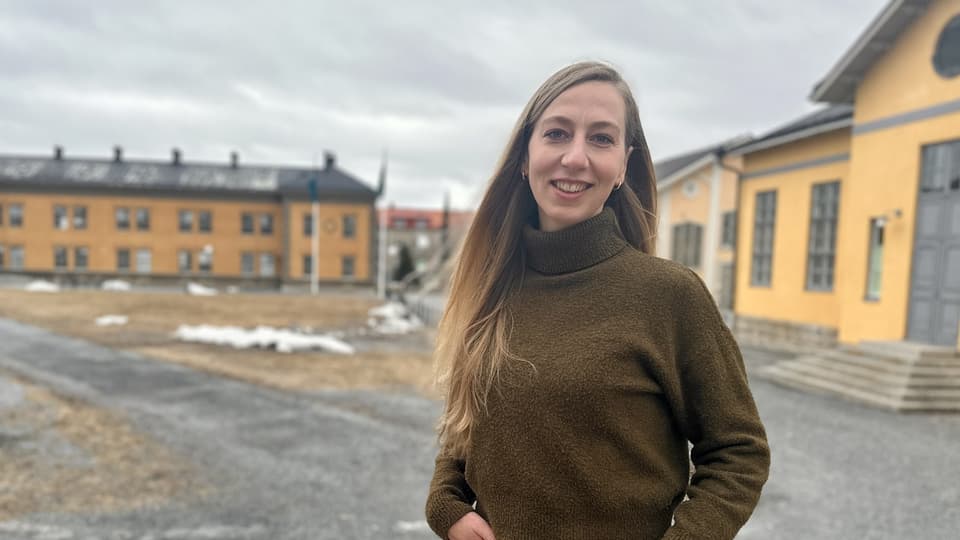 Woman in brown polo shirt and black trousers standing outdoors in front of large yellow buildings