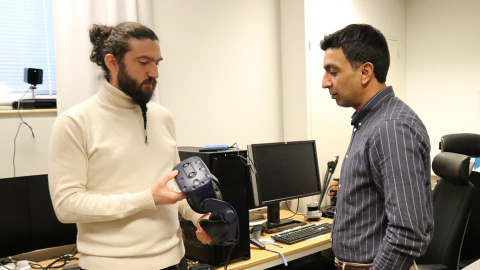  Two men stand together in a computer room. One man is holding a pair of VR glasses.