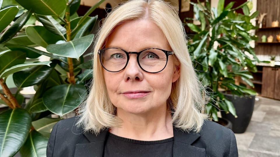 En kvinna med axellångt blont hår och glasögon, klädd i svart kavaj står framför gröna, höga växter i inomhusmiljö. 