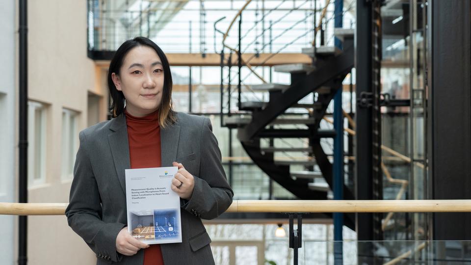 A woman indoors holding a thesis with both hands and smiling at the camera