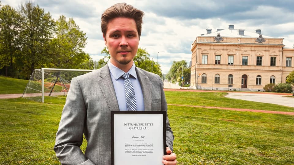 En man i grå kostym håller i ett diplom. I bakgrunden finns en gräsmatta och en byggnad.