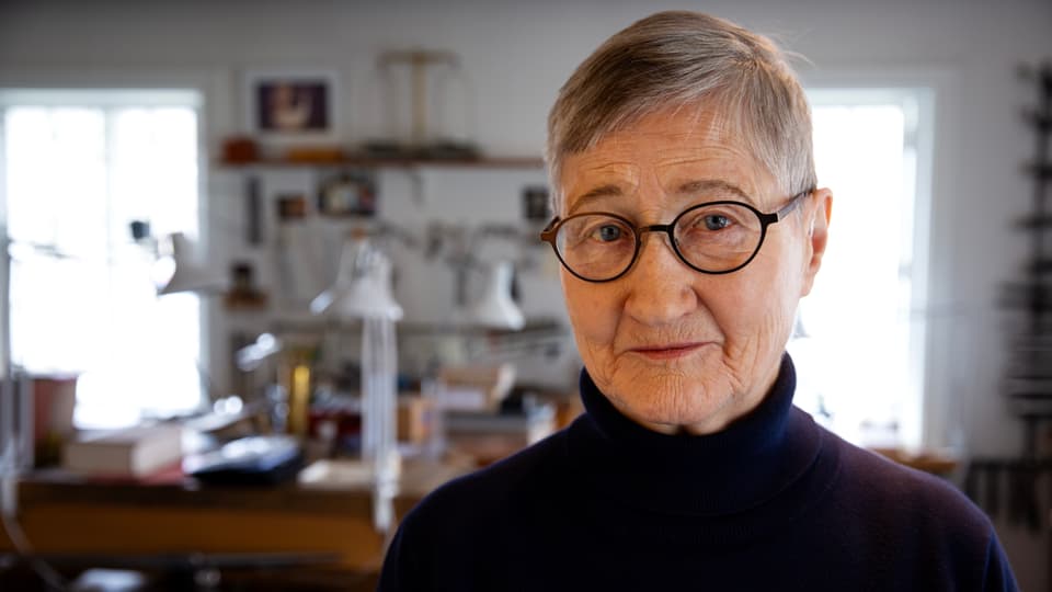 A woman in glasses and short hair is looking into the camera. In the background you can see desks and shelfs.