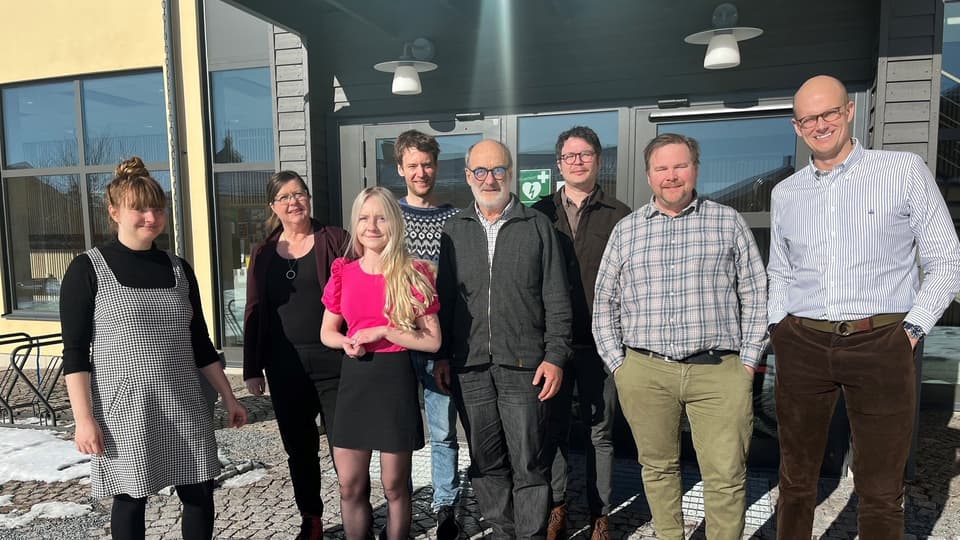 En grupp människor står framför entrén till en gul byggnad. Solen skiner.