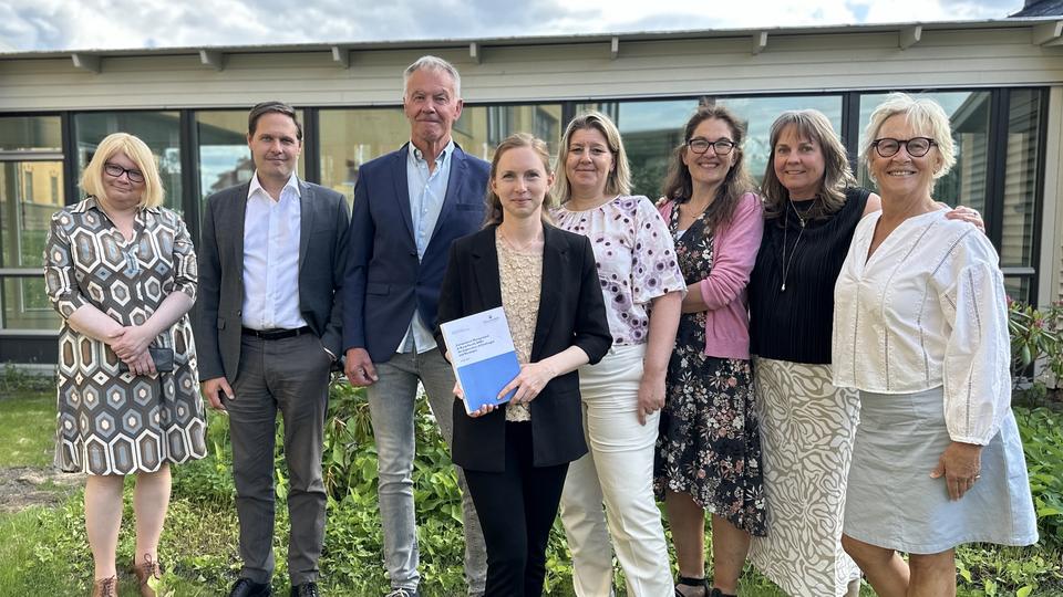 Åtta personer, två män och sex kvinnor står tillsammans i utemiljö och en kvinna står längst fram och håller i en avhandling