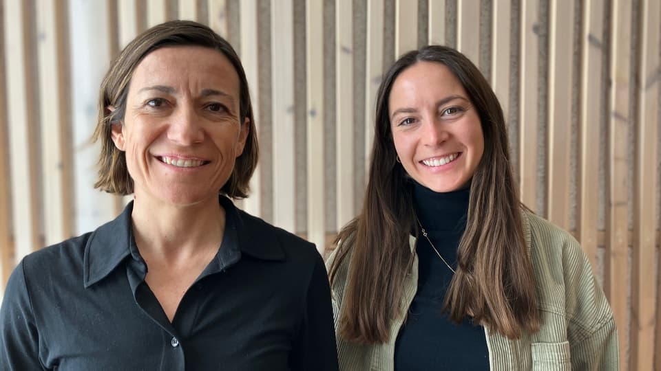 Two women stand in front of a wood-panelled wall and look into the camera, smiling