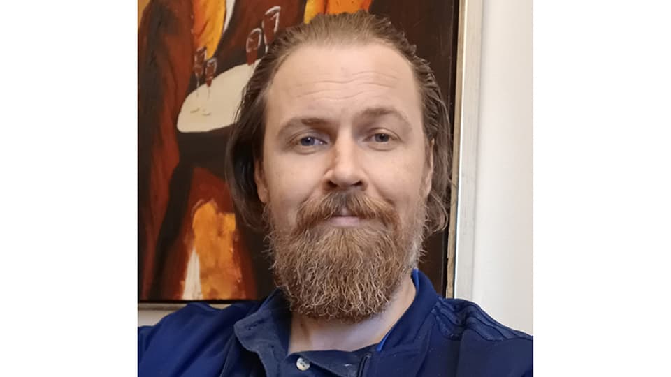 Man med rödlätt hår och skägg, klädd i en blå skjorta.