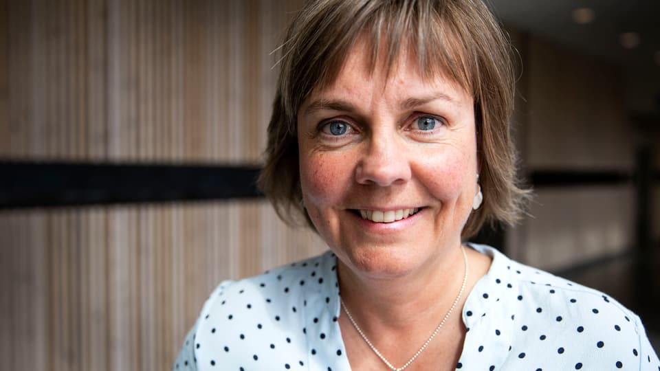 Kvinna med kortklippt frisyr och prickig blus står framför en vägg med träpanel och ler.  