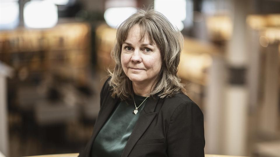 En kvinna i ljusbrunt hår, grön blus och svart kavaj står i biblioteksmiljö.