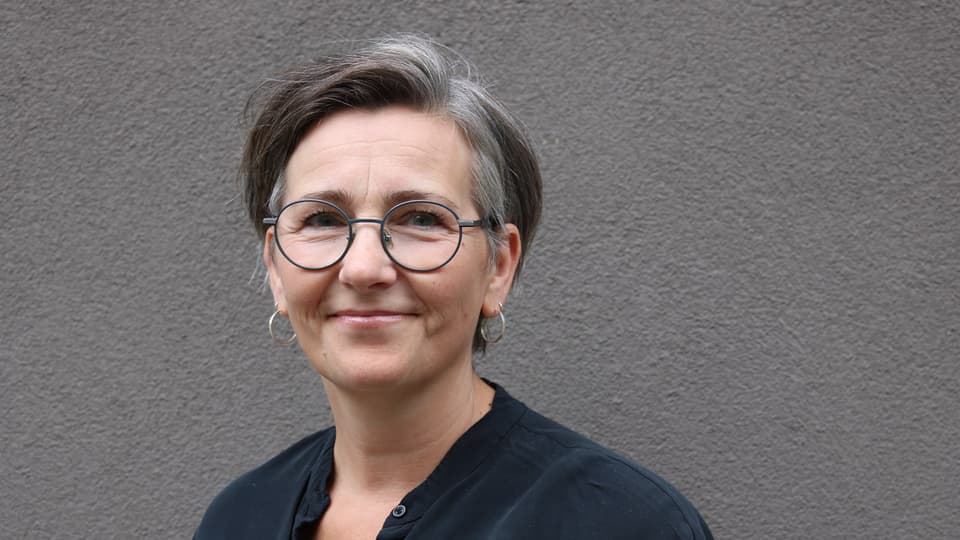 En kvinna med mörkt hår, glasögon och en svart blus står framför en mörkgrå vägg.