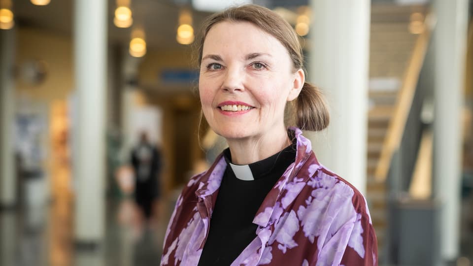 Kvinna med svart skjorta med prästkrage ler mot kameran. 