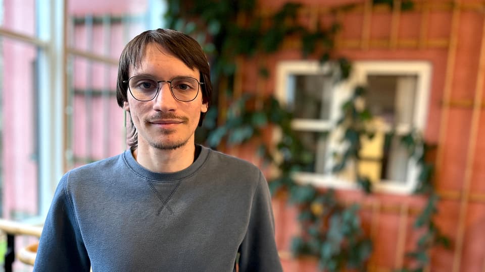 A man with brown hair, glasses and a grey jumper stands in front of a reddish-brown wall covered in climbing green plants. 