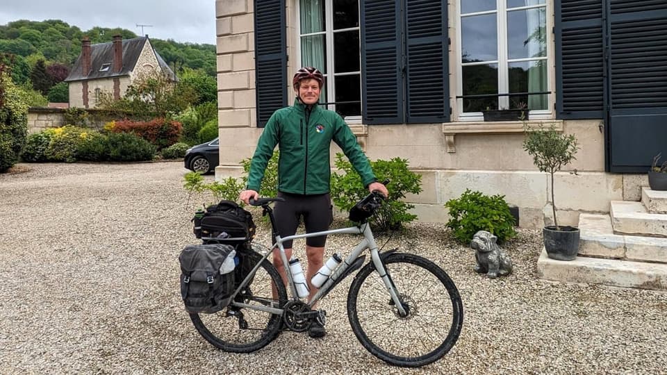 En man i grön jacka och röd cykelhjälm står framför ett hus med sin cykel framför sig