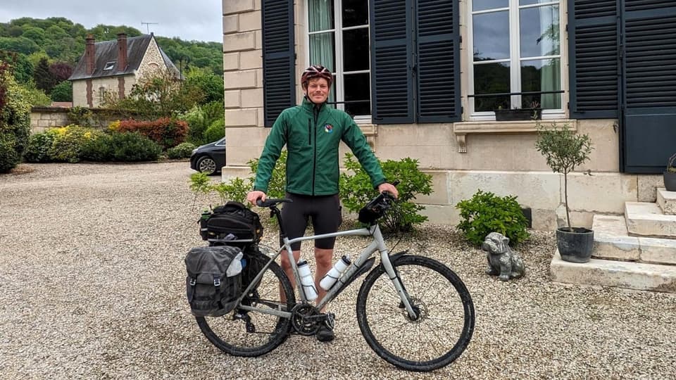 En man i grön jacka och röd cykelhjälm står framför ett hus med sin cykel framför sig
