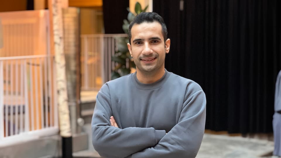  A man with dark hair and a gray sweatshirt stands with his arms crossed, smiling at the camera.