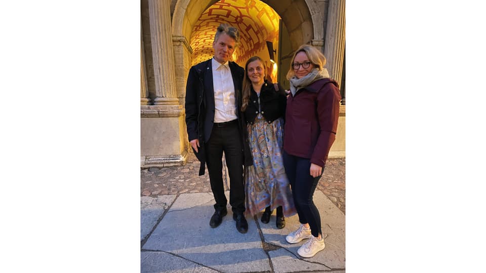 A man and two women stand in front of an illuminated stone archway.