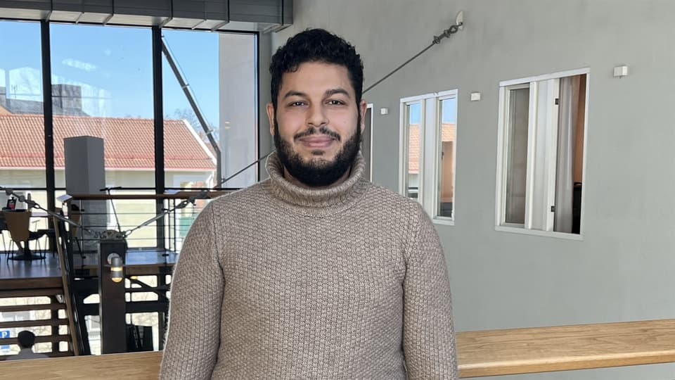  A young man in a beige sweater looks into the camera and smiles.