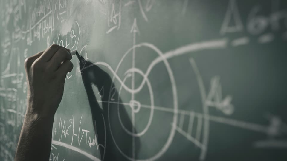  A hand holds a white chalk and writes formulas on a blackboard.