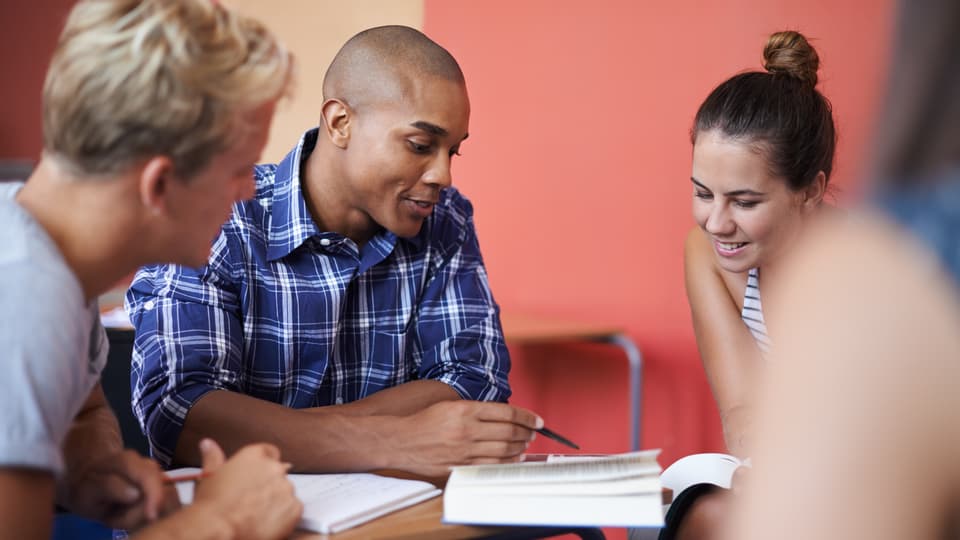 Grupp bestående av fyra studenter sitter och diskuterar runt en bok. Bakom dem syns en röd vägg och de ser glada ut.