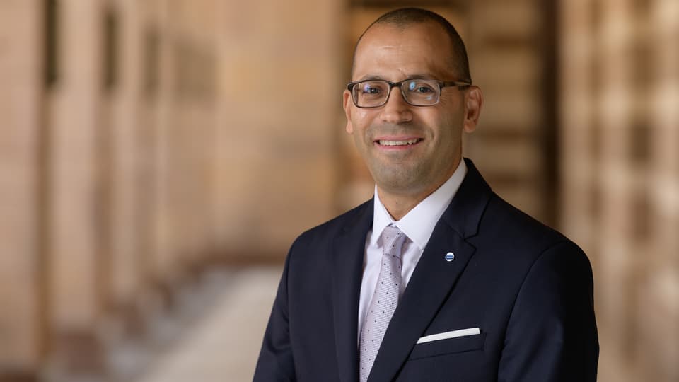 A well-dressed man with short hair and glasses smiles at the camera.