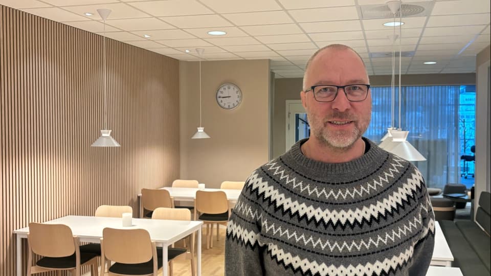 Man i grå stickad tröja med glasögon ler mot kameran, en sal med ljust interiör, stolar och vita bord i bakgrunden.