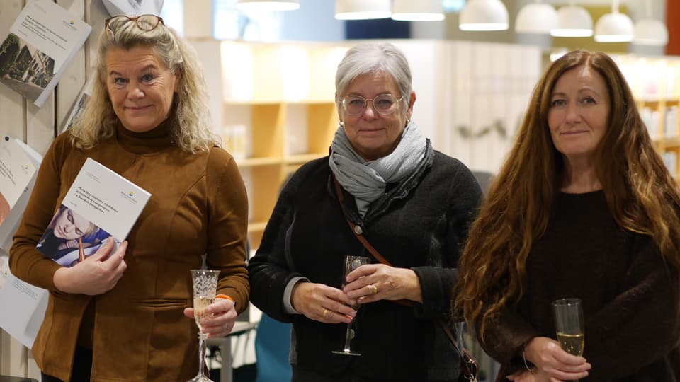 Tre kvinnor står bredvid varandra med champagneglas i händerna. Kvinnan längst till vänster håller upp en bok.