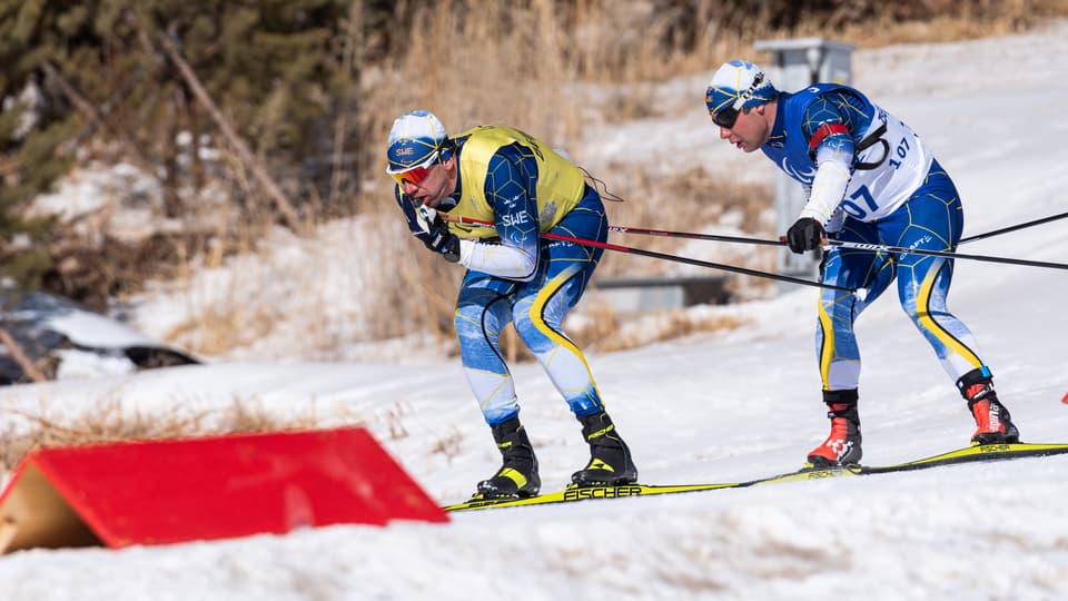 Två skidåkare i blågula dräkter som åker utför en bana märkt med röda v-boards. Den bakre skidåkaren håller i den främre åkarens ena skidstav.