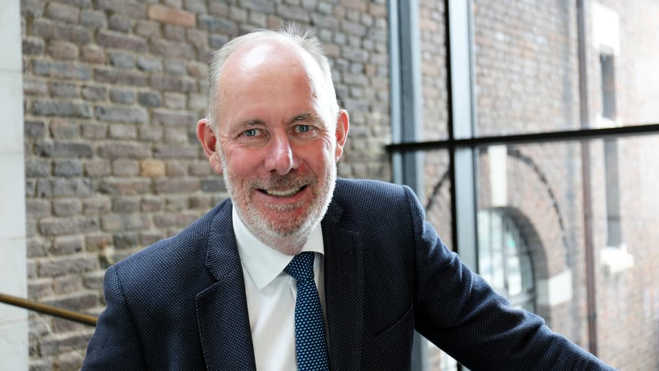 Man in dark blue jacket, white shirt and tie smiles and looks at the camera.