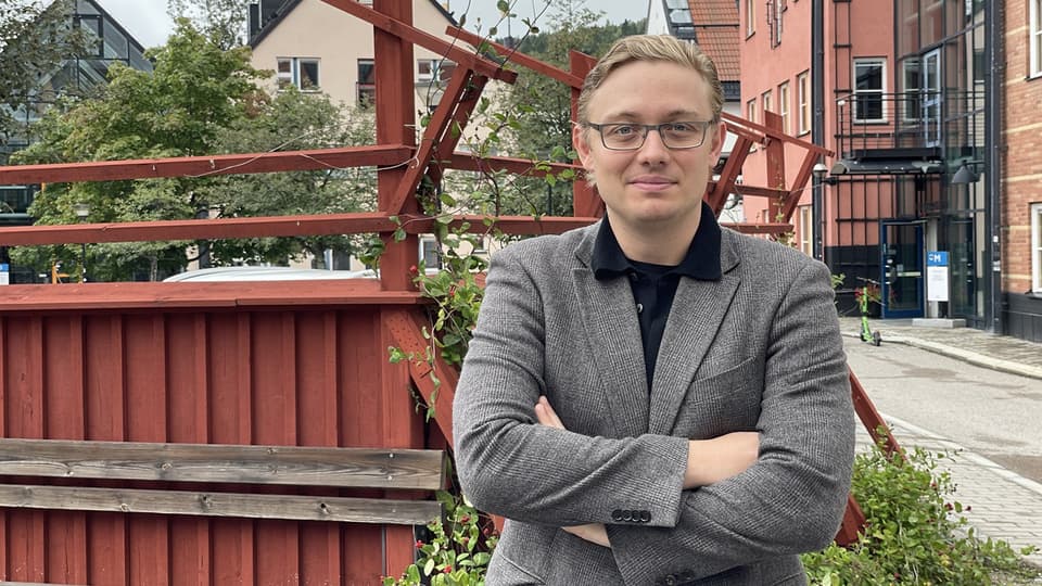 A fair-haired man with glasses and a blazer stands in front of a red fence and poses for the camera.
