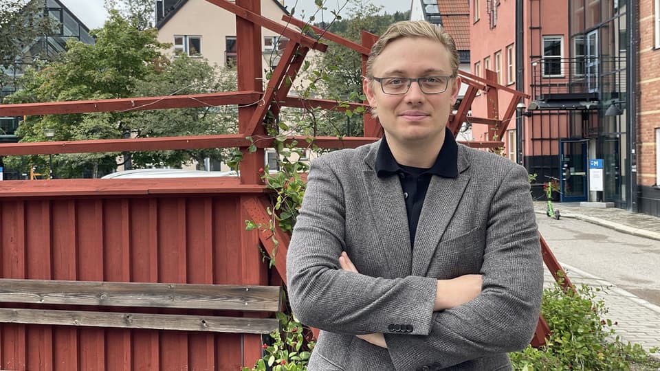 A fair-haired man with glasses and a blazer stands in front of a red fence and poses for the camera.