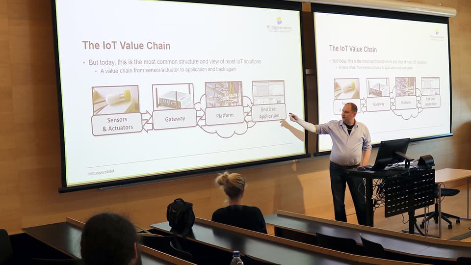 A man in a light blue shirt stands in front of a large screen and gives a presentation.